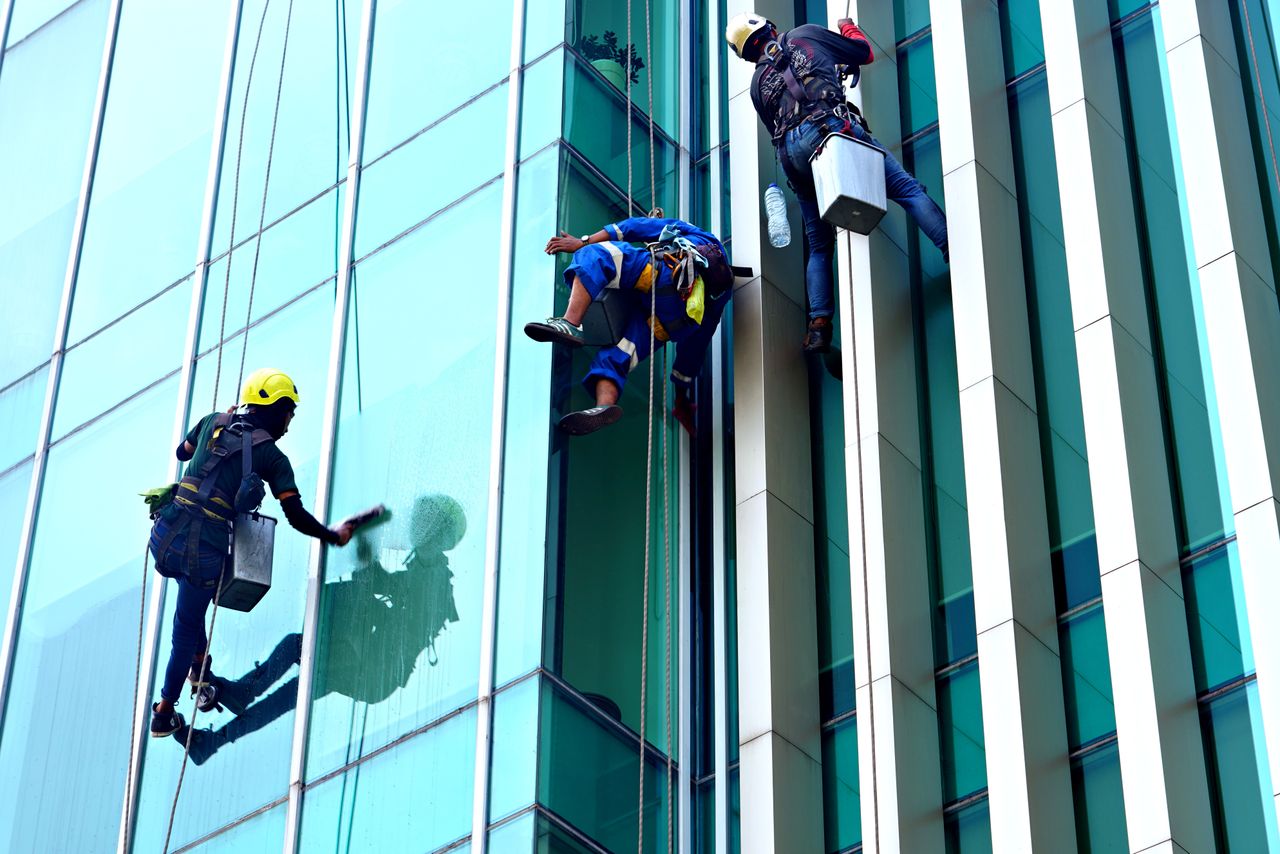 Three window cleaners suspended on ropes clean a modern glass building facade during the day.